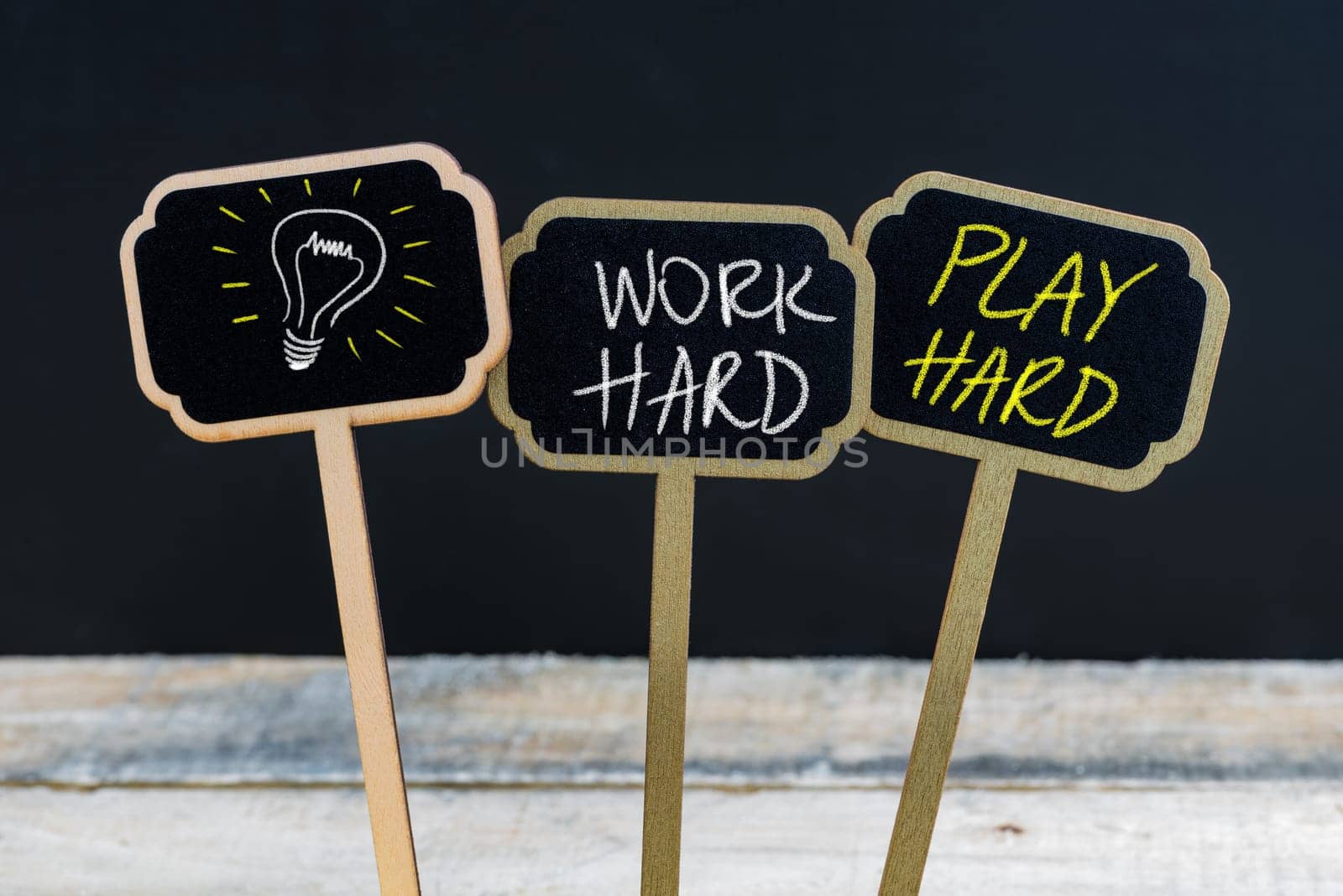 Concept message WORK HARD PLAY HARD and light bulb as symbol for idea written with chalk on wooden mini blackboard labels, defocused chalkboard and wood table in background