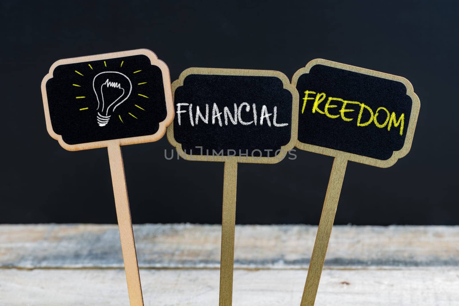 Concept message FINANCIAL FREEDOM and light bulb as symbol for idea written with chalk on wooden mini blackboard labels, defocused chalkboard and wood table in background