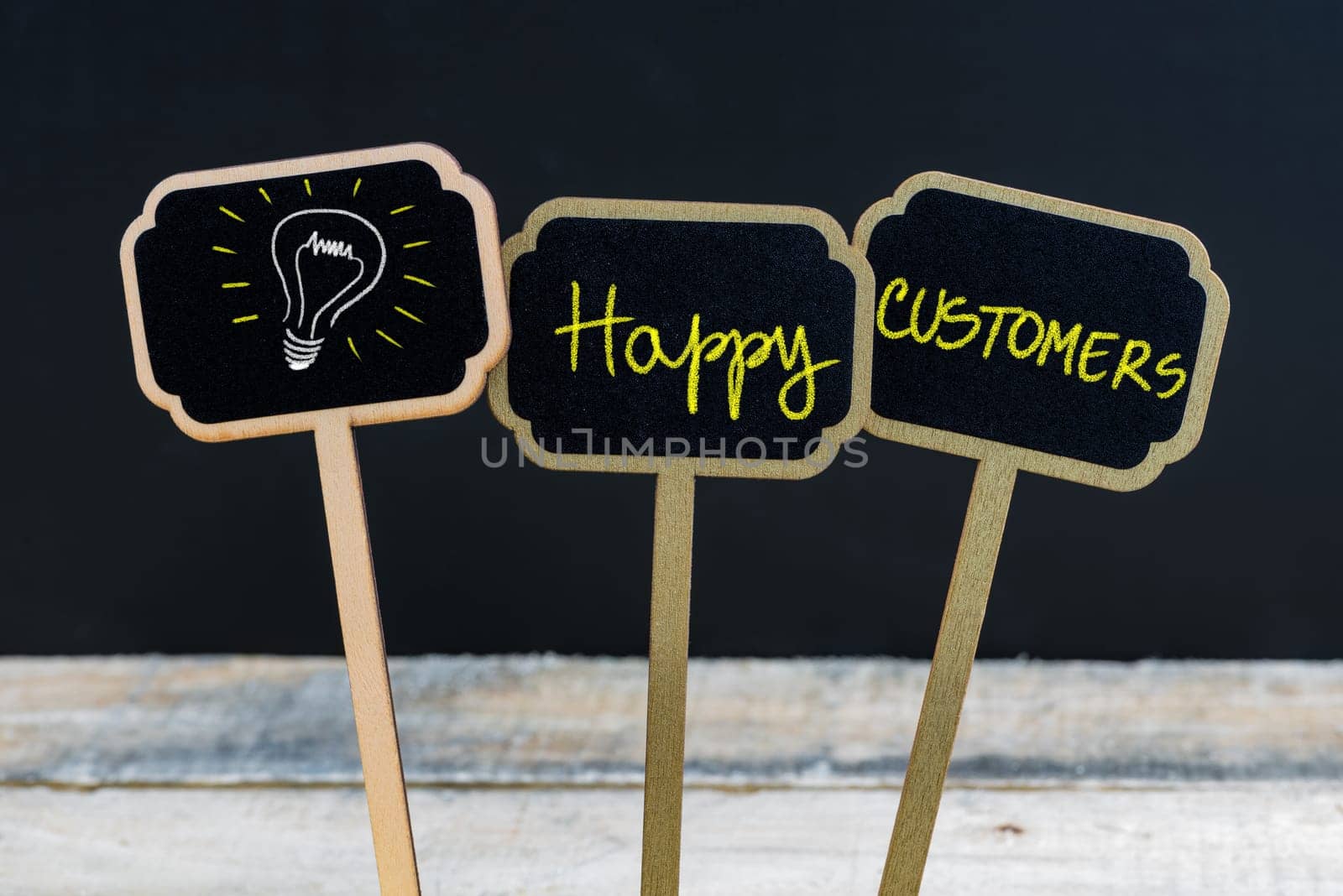 Concept message Happy Customers and light bulb as symbol for idea written with chalk on wooden mini blackboard labels, defocused chalkboard and wood table in background