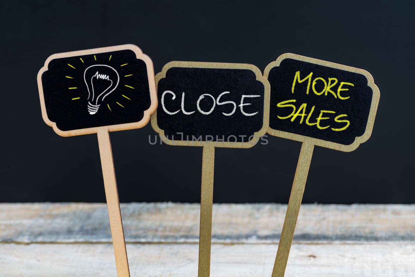 Concept message CLOSE MORE SALES and light bulb as symbol for idea written with chalk on wooden mini blackboard labels, defocused chalkboard and wood table in background