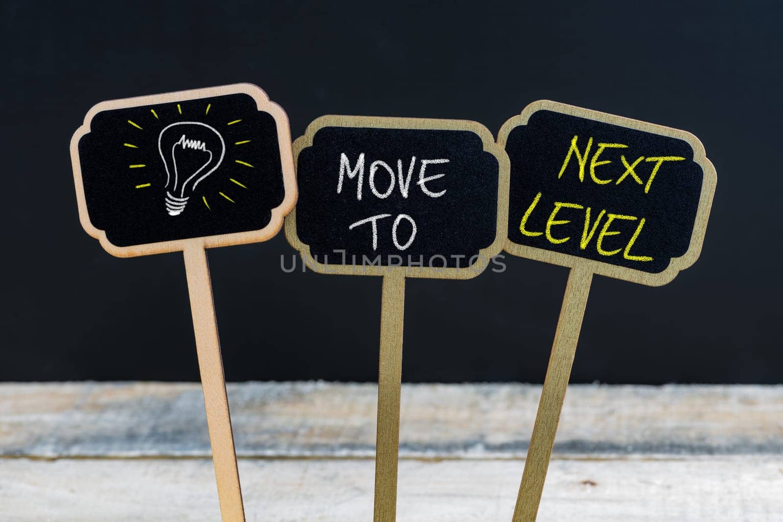 Concept message MOVE TO NEXT LEVEL and light bulb as symbol for idea written with chalk on wooden mini blackboard labels, defocused chalkboard and wood table in background