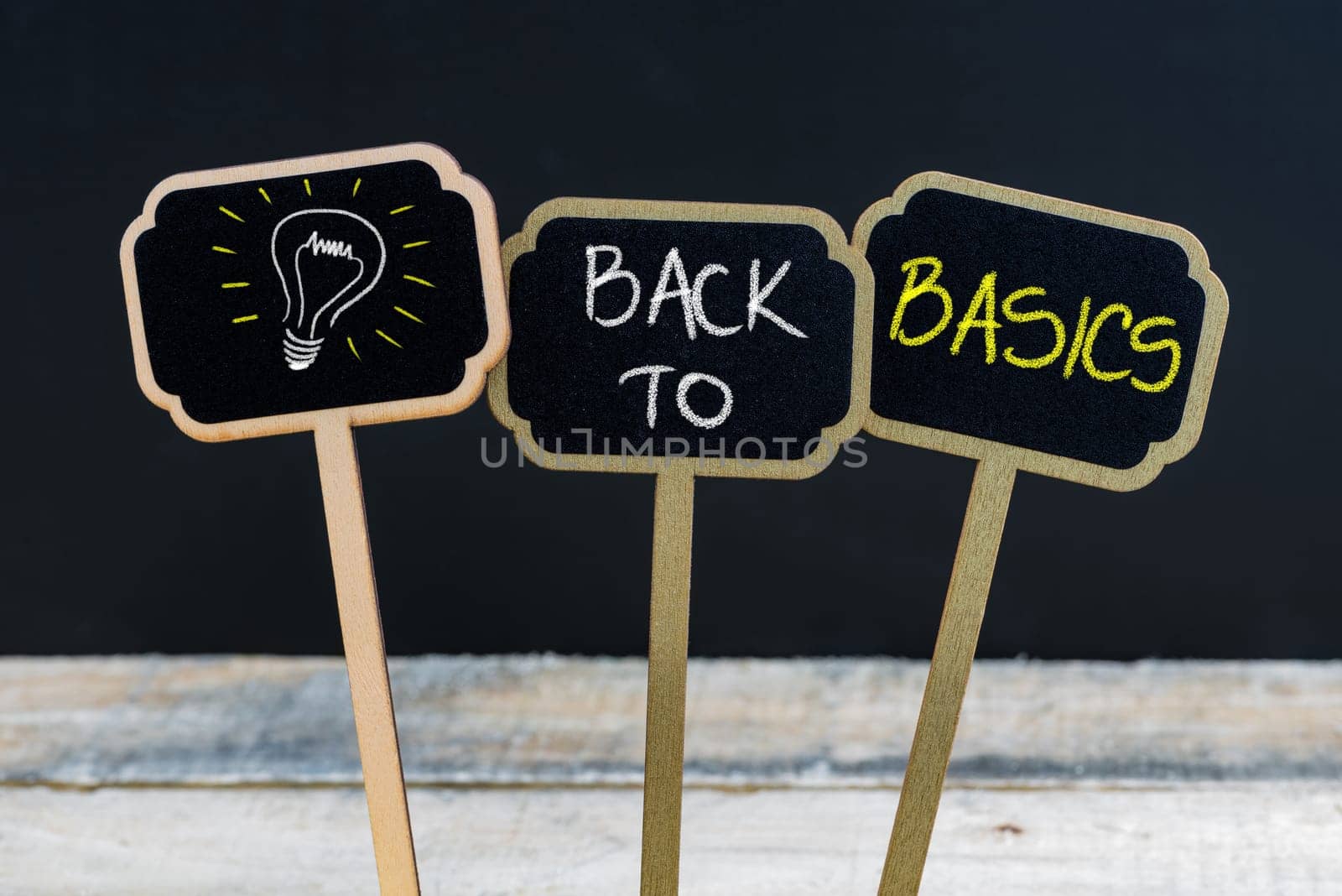 Concept message BACK TO BASICS and light bulb as symbol for idea written with chalk on wooden mini blackboard labels, defocused chalkboard and wood table in background