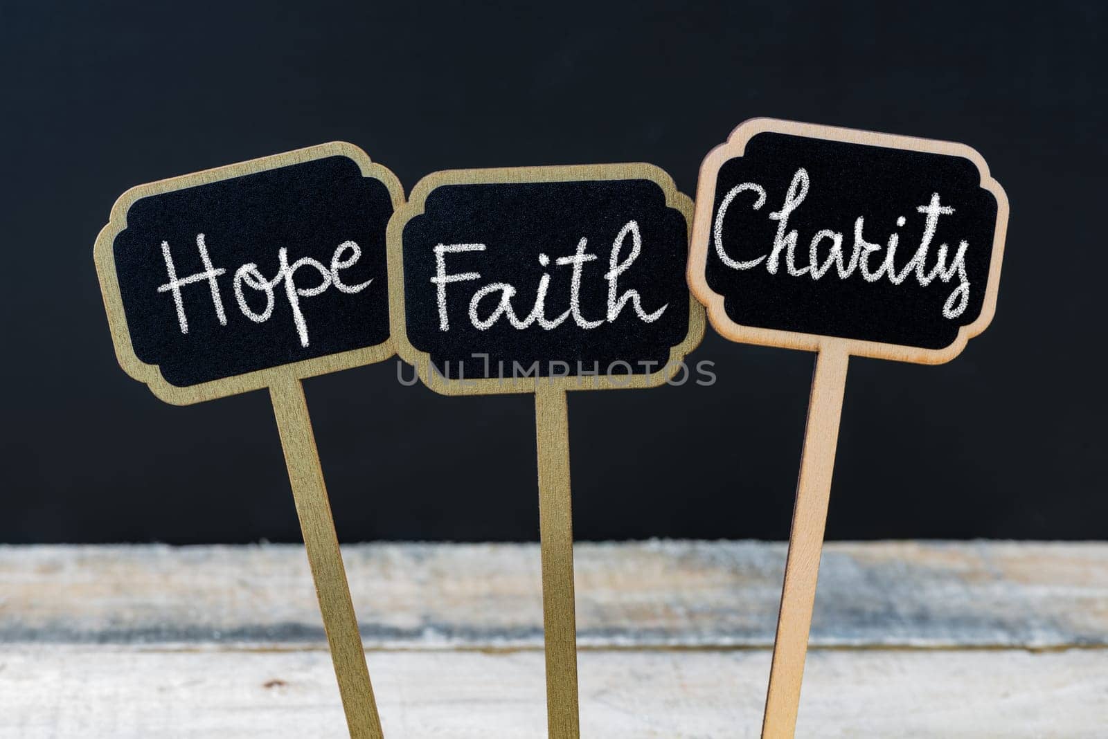 Concept message Hope Faith Charity written with chalk on wooden mini blackboard labels, defocused chalkboard and wood table in background