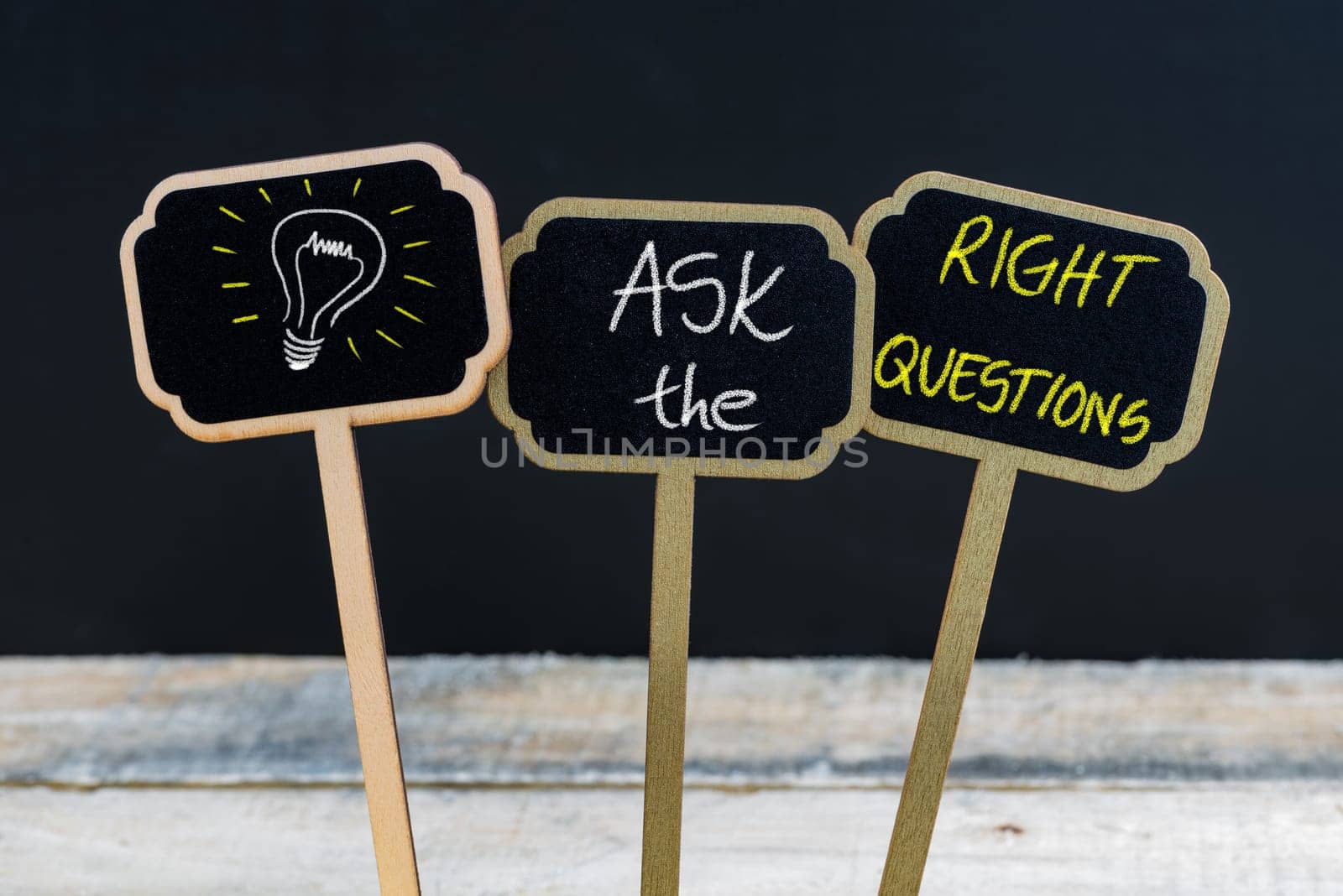 Concept message ASK THE RIGHT QUESTIONS and light bulb as symbol for idea written with chalk on wooden mini blackboard labels, defocused chalkboard and wood table in background