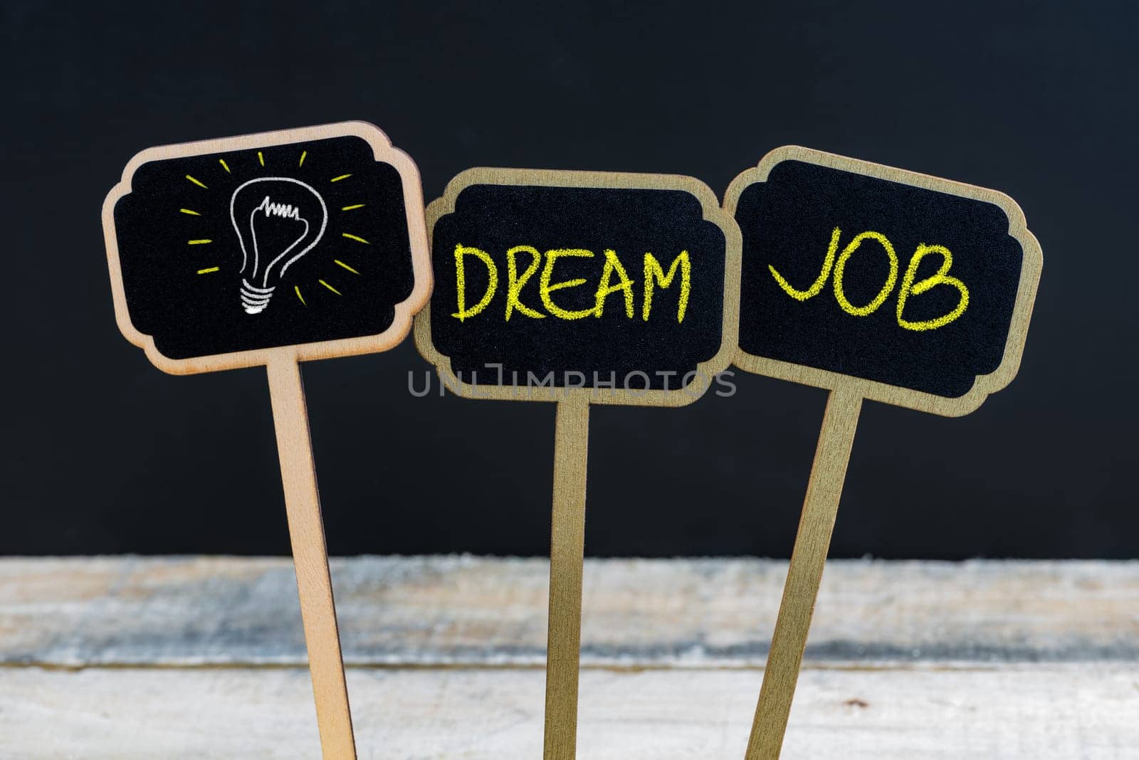 Concept message DREAM JOB and light bulb as symbol for idea written with chalk on wooden mini blackboard labels, defocused chalkboard and wood table in background