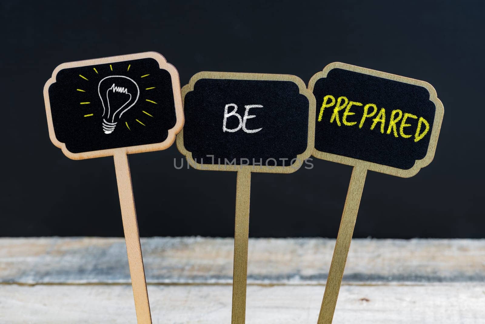 Concept message BE PREPARED and light bulb as symbol for idea written with chalk on wooden mini blackboard labels, defocused chalkboard and wood table in background
