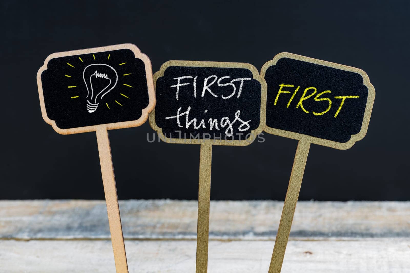Concept message FIRST THINGS FIRST and light bulb as symbol for idea written with chalk on wooden mini blackboard labels, defocused chalkboard and wood table in background