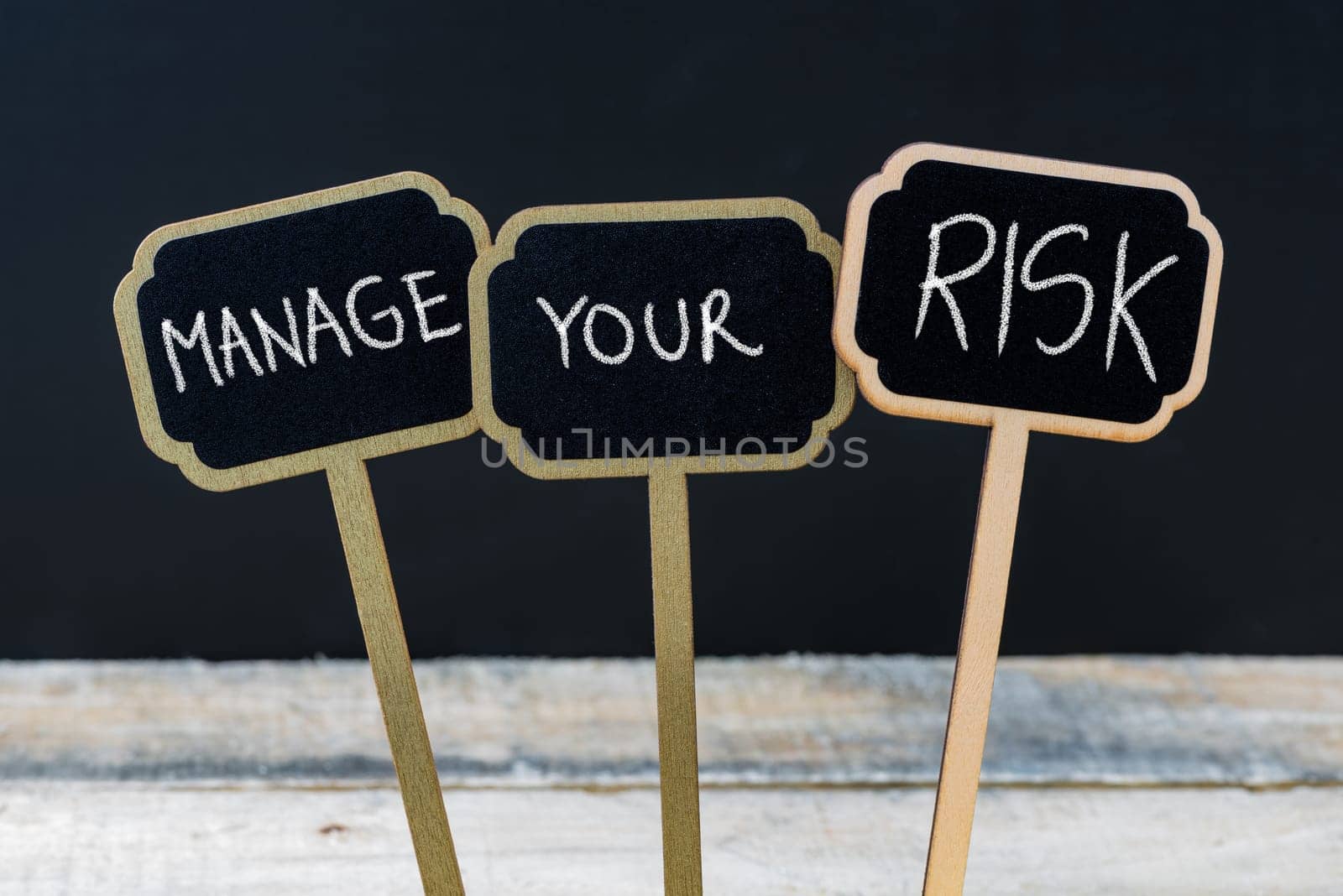 Concept message MANAGE YOUR RISK written with chalk on wooden mini blackboard labels, defocused chalkboard and wood table in background