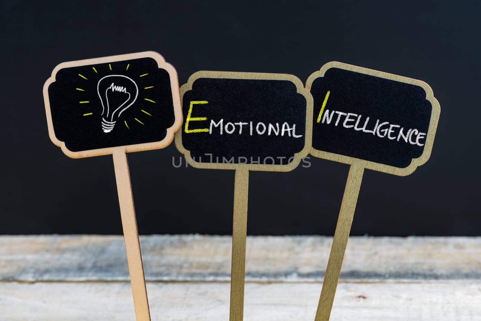 Concept message EMOTIONAL INTELLIGENCE and light bulb as symbol for idea written with chalk on wooden mini blackboard labels, defocused chalkboard and wood table in background