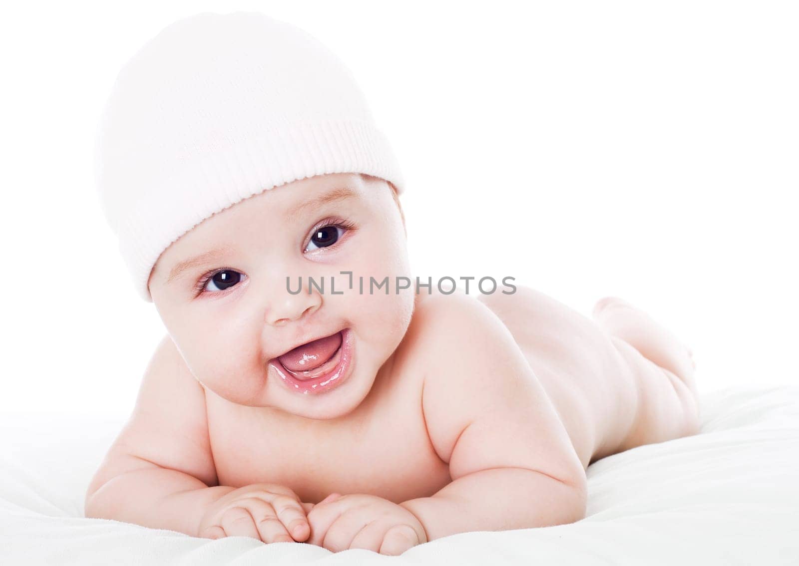 Happy baby lying on stomach on white floor background, wearing a cap and smiling big.