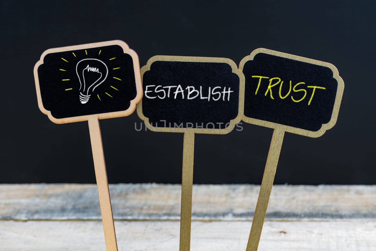 Concept message ESTABLISH TRUST and light bulb as symbol for idea written with chalk on wooden mini blackboard labels, defocused chalkboard and wood table in background