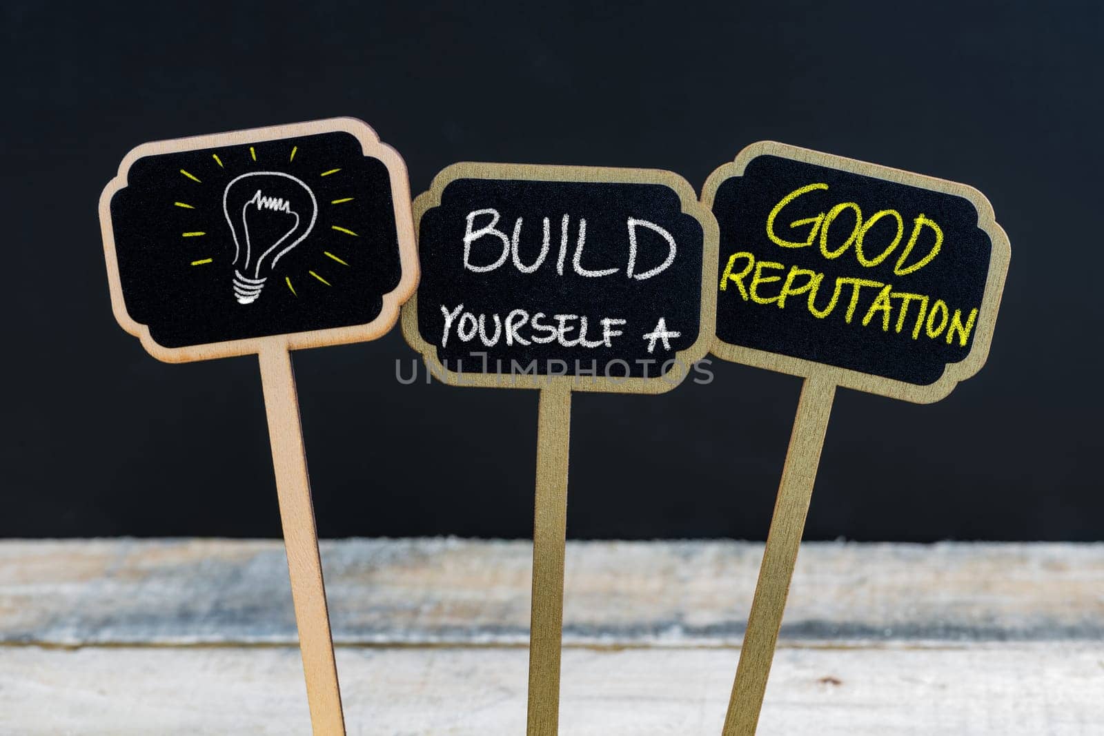 Concept message BUILD YOURSELF A GOOD REPUTATION and light bulb as symbol for idea written with chalk on wooden mini blackboard labels, defocused chalkboard and wood table in background