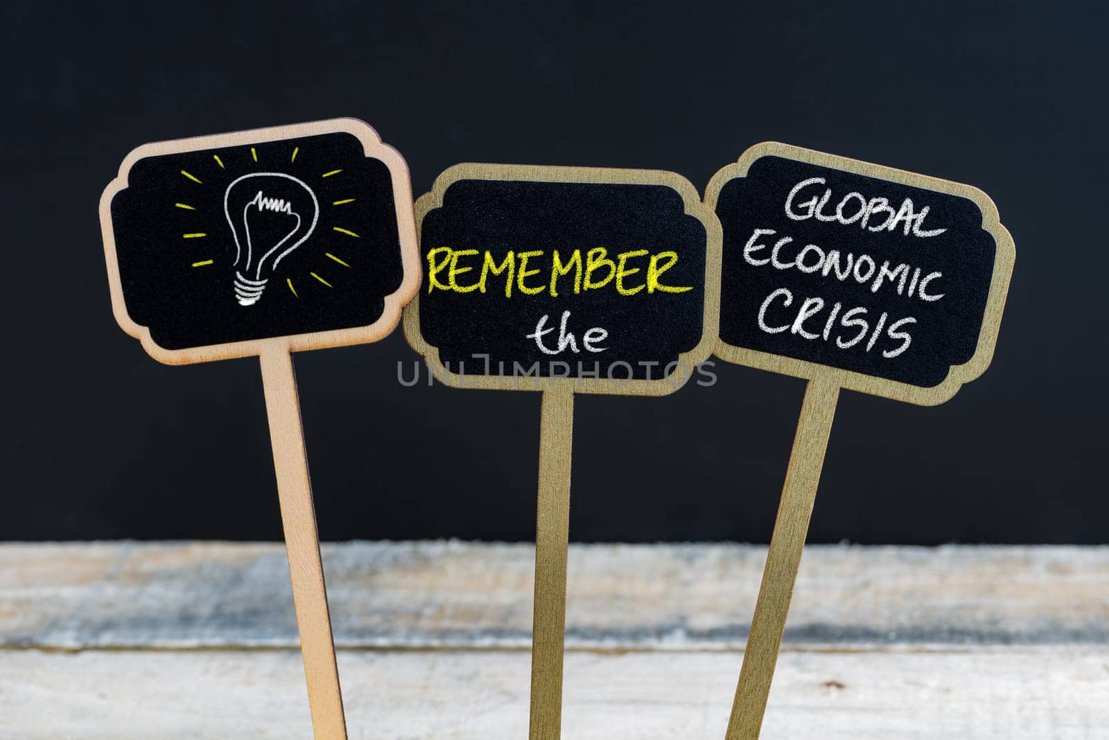 Concept message REMEMBER THE GLOBAL ECONOMIC CRISIS and light bulb as symbol for idea written with chalk on wooden mini blackboard labels, defocused chalkboard and wood table in background