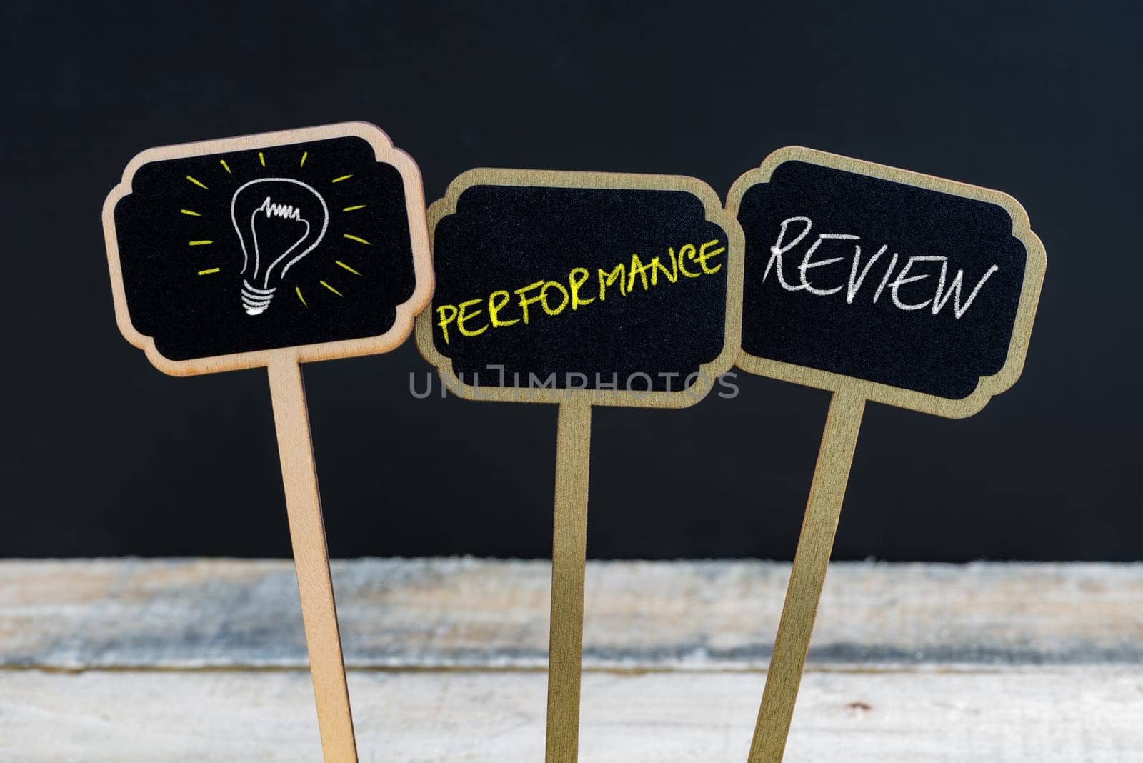 Concept message PERFORMANCE REVIEW and light bulb as symbol for idea written with chalk on wooden mini blackboard labels, defocused chalkboard and wood table in background