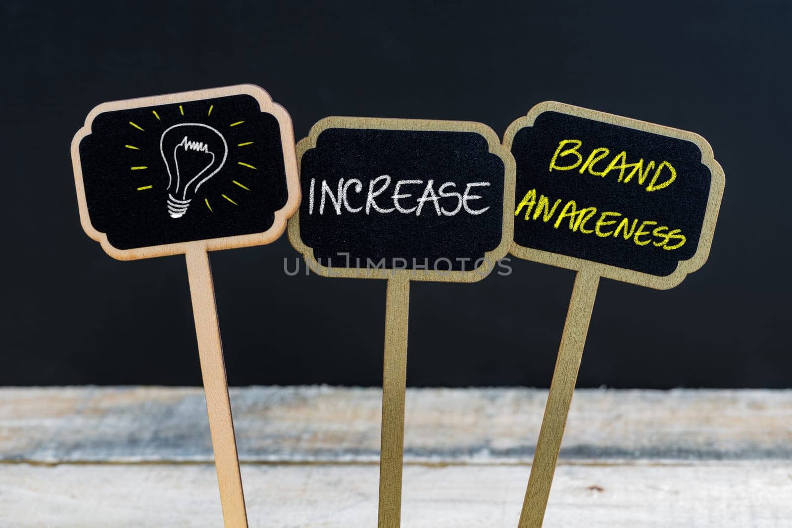 Concept message INCREASE BRAND AWARENESS and light bulb as symbol for idea written with chalk on wooden mini blackboard labels, defocused chalkboard and wood table in background