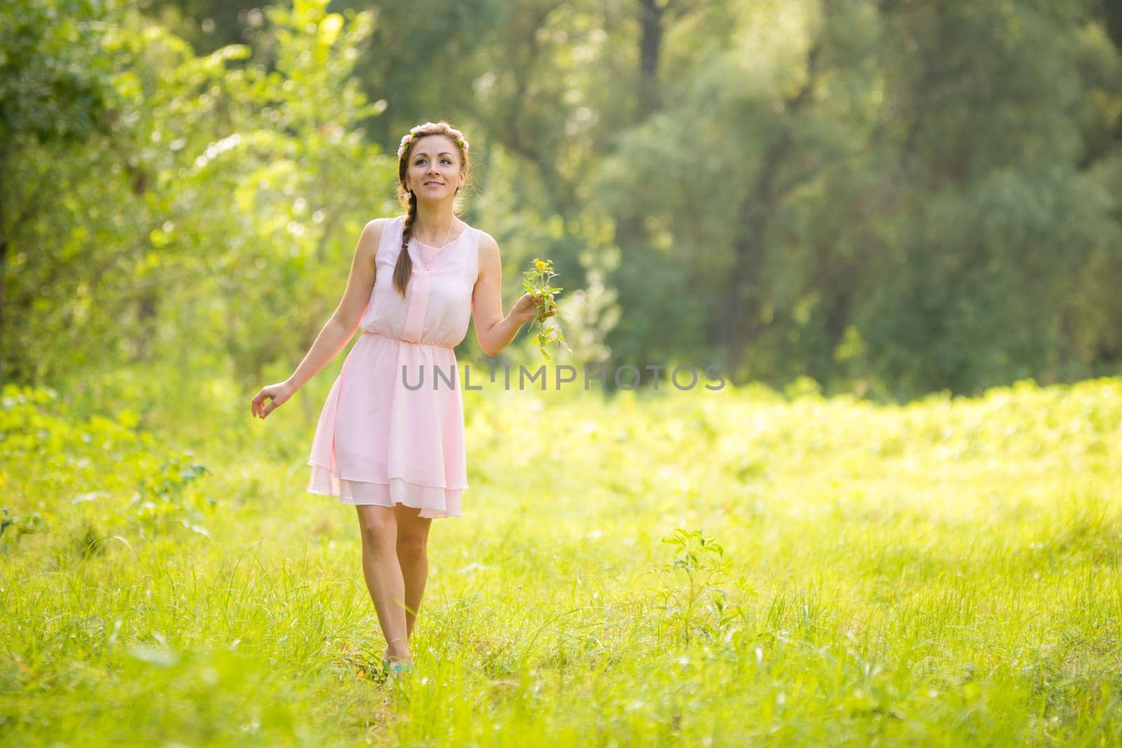 Young beautiful woman walking on a green meadow with a bouquet of wildflowers in a light summer dress