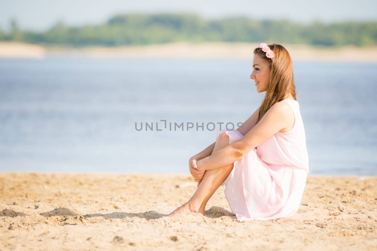 Young girl in the summer light white dress sitting on the sandy beach by the river