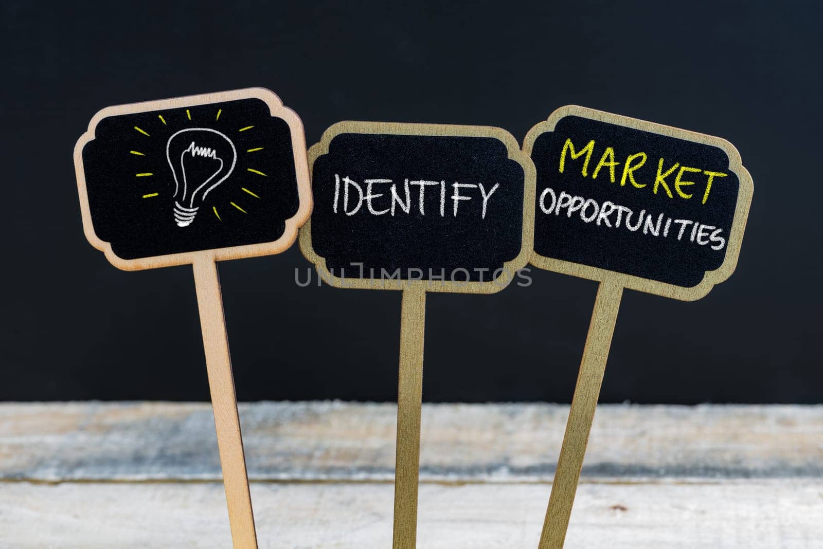 Concept message IDENTIFY MARKET OPPORTUNITIES and light bulb as symbol for idea written with chalk on wooden mini blackboard labels, defocused chalkboard and wood table in background