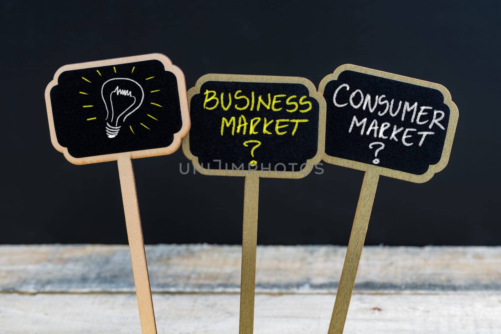 Concept message BUSINESS MARKET versus CONSUMER MARKET and light bulb as symbol for idea written with chalk on wooden mini blackboard labels, defocused chalkboard and wood table in background