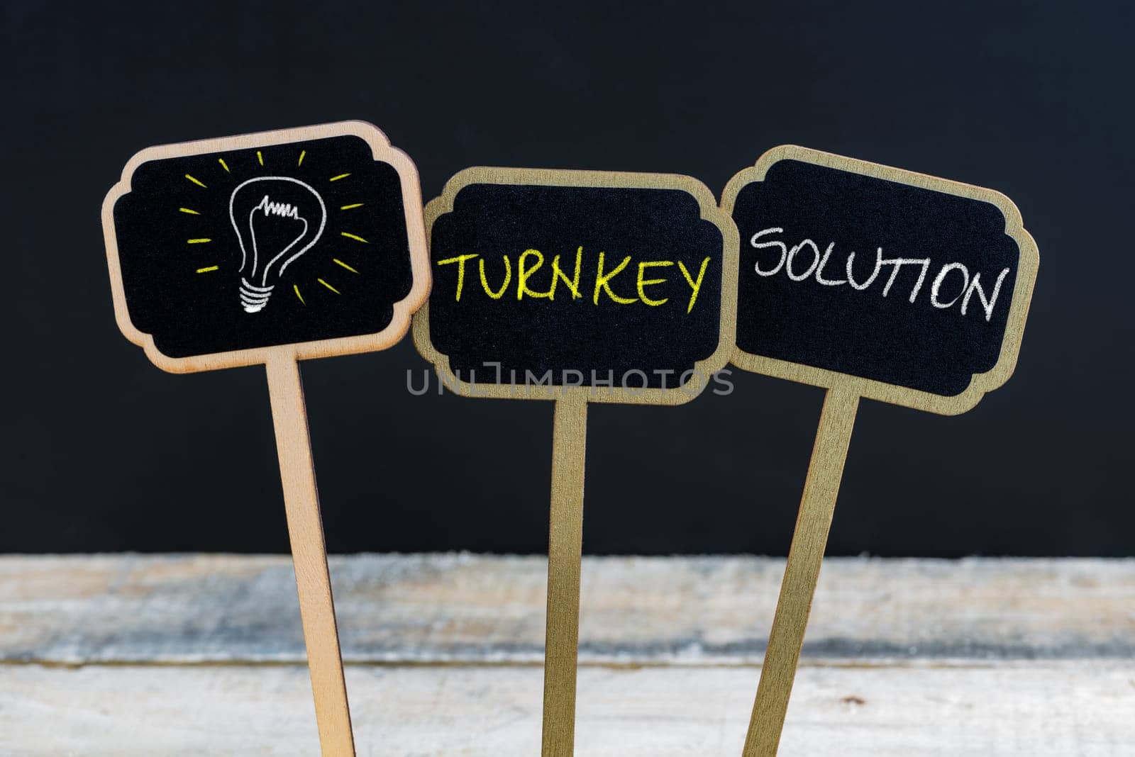 Concept message TURNKEY SOLUTION and light bulb as symbol for idea written with chalk on wooden mini blackboard labels, defocused chalkboard and wood table in background