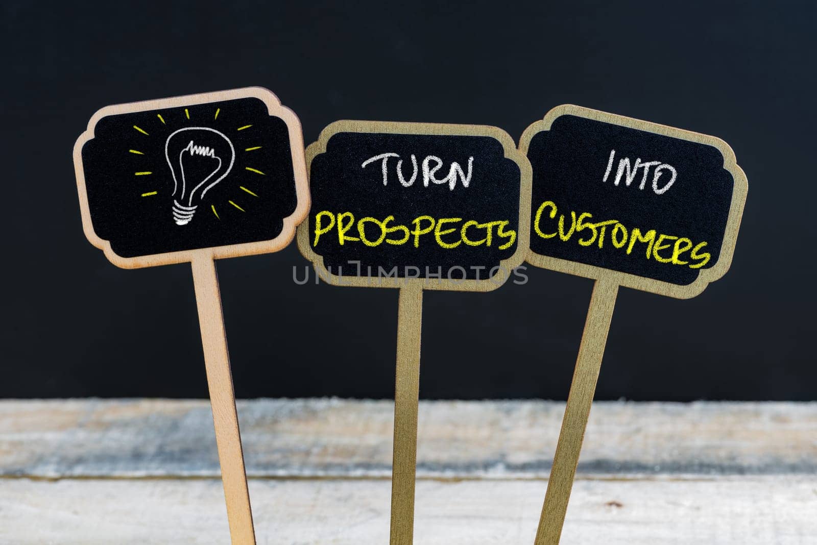 Concept message TURN PROSPECTS INTO CUSTOMERS and light bulb as symbol for idea written with chalk on wooden mini blackboard labels, defocused chalkboard and wood table in background