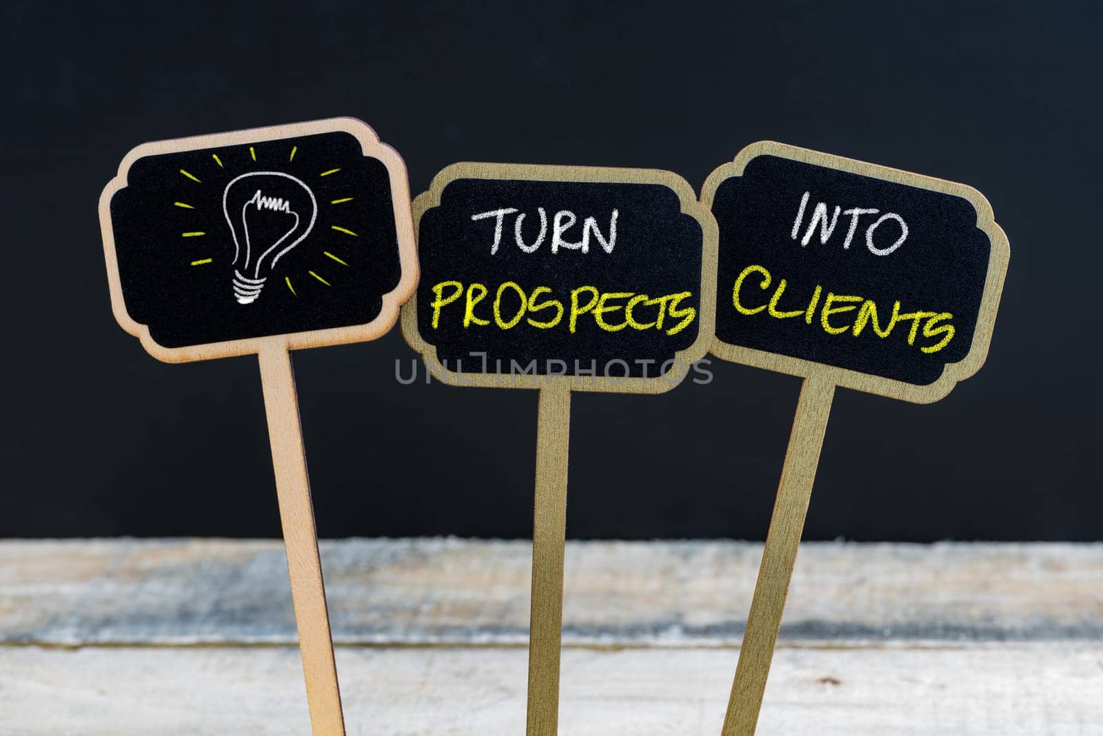 Concept message TURN PROSPECTS INTO CLIENTS and light bulb as symbol for idea written with chalk on wooden mini blackboard labels, defocused chalkboard and wood table in background