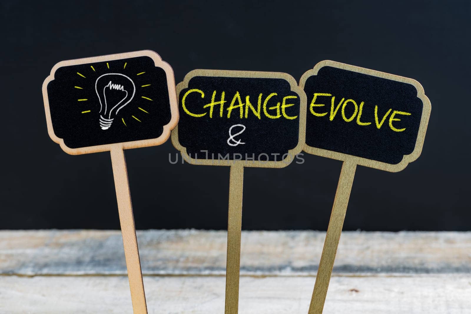 Concept message CHANGE AND EVOLVE and light bulb as symbol for idea written with chalk on wooden mini blackboard labels, defocused chalkboard and wood table in background