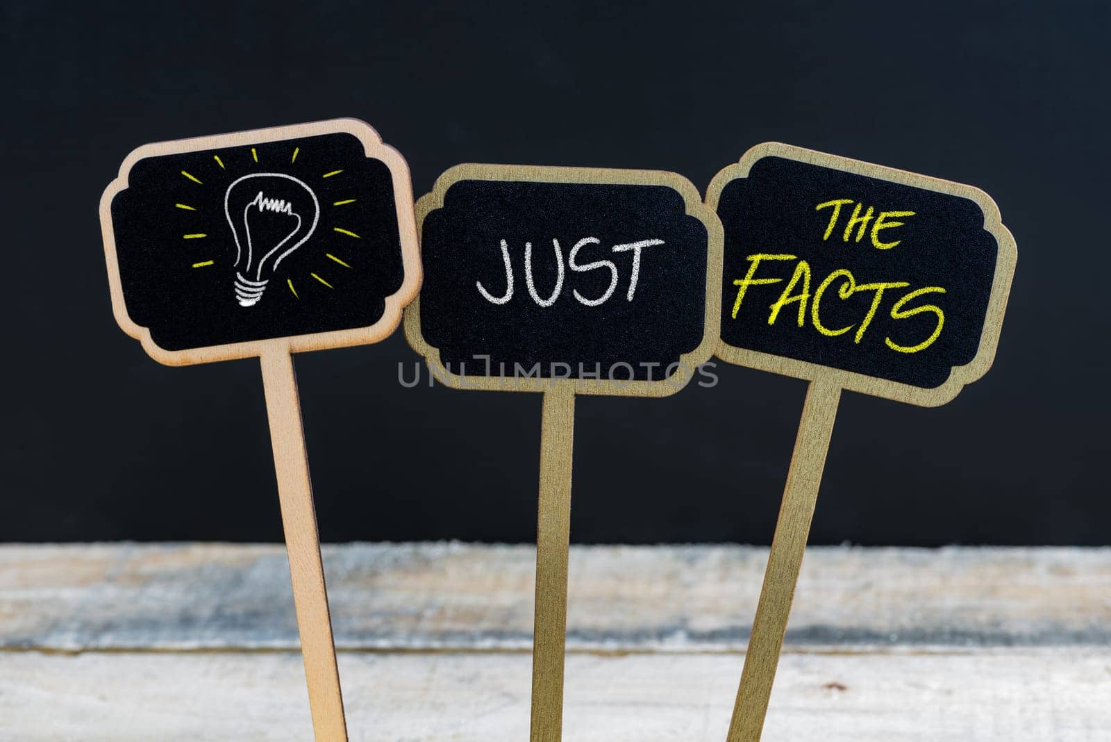 Concept message JUST THE FACTS and light bulb as symbol for idea written with chalk on wooden mini blackboard labels, defocused chalkboard and wood table in background