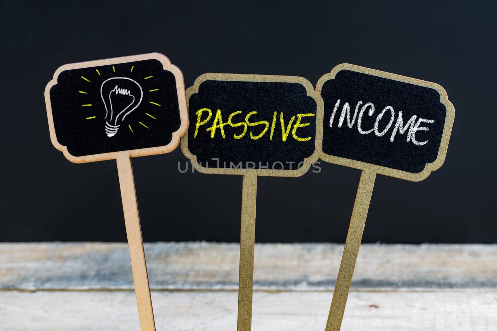 Concept message PASSIVE INCOME and light bulb as symbol for idea written with chalk on wooden mini blackboard labels, defocused chalkboard and wood table in background