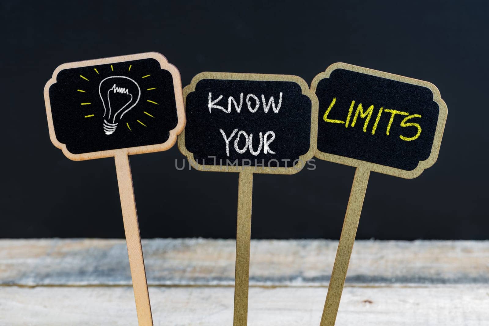 Concept message KNOW YOUR LIMITS and light bulb as symbol for idea written with chalk on wooden mini blackboard labels, defocused chalkboard and wood table in background