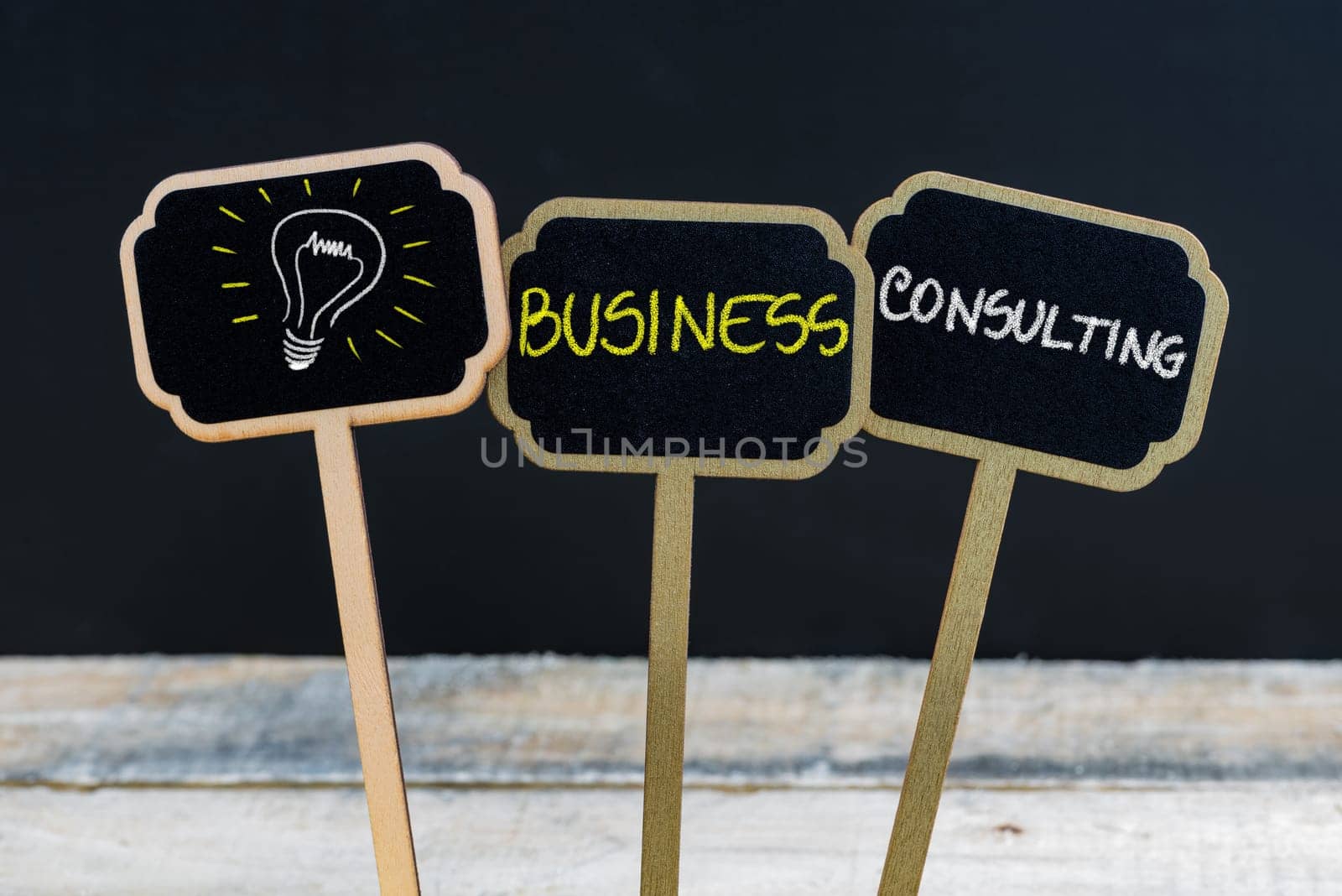 Concept message BUSINESS CONSULTING and light bulb as symbol for idea written with chalk on wooden mini blackboard labels, defocused chalkboard and wood table in background