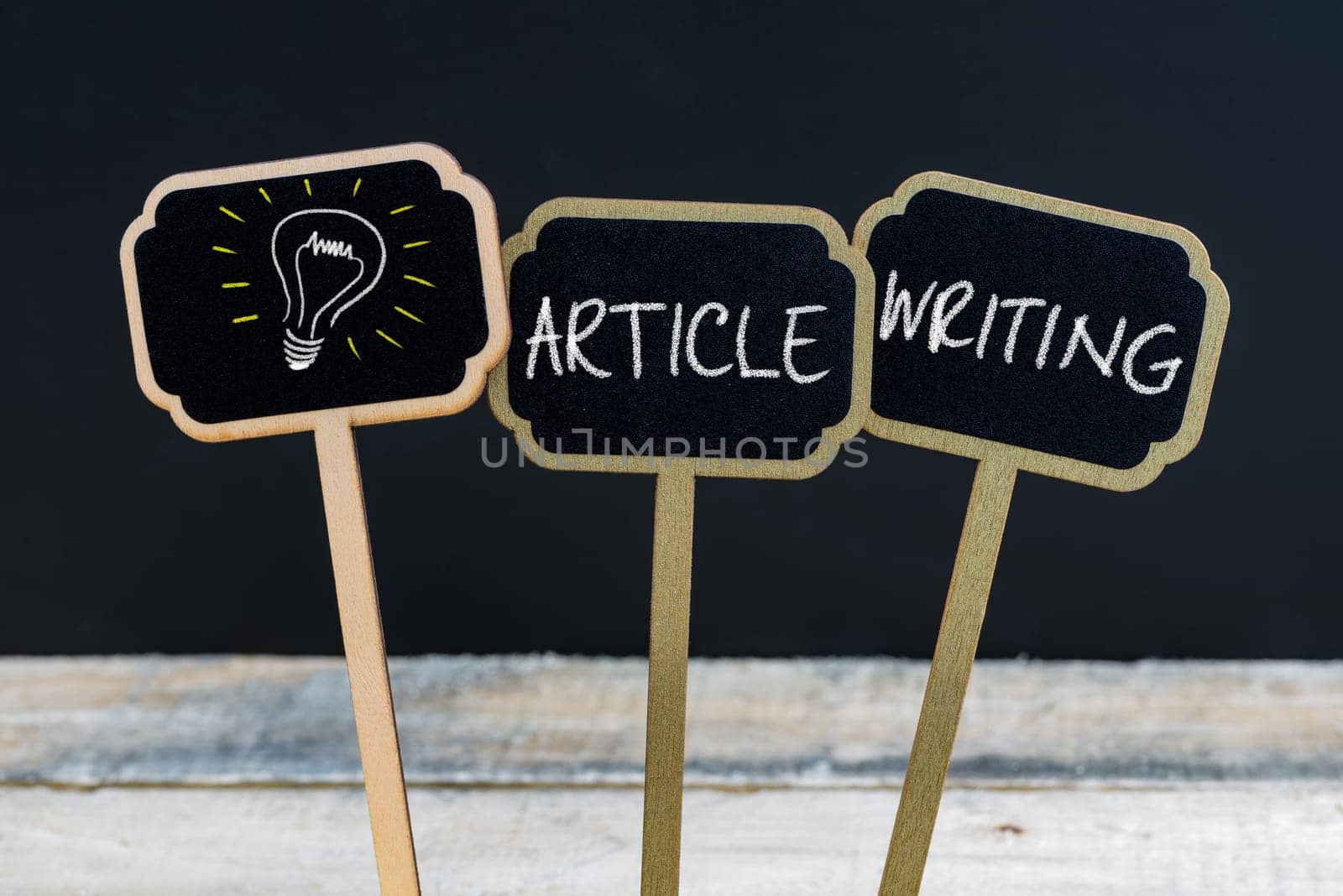 Concept message ARTICLE WRITING and light bulb as symbol for idea written with chalk on wooden mini blackboard labels, defocused chalkboard and wood table in background