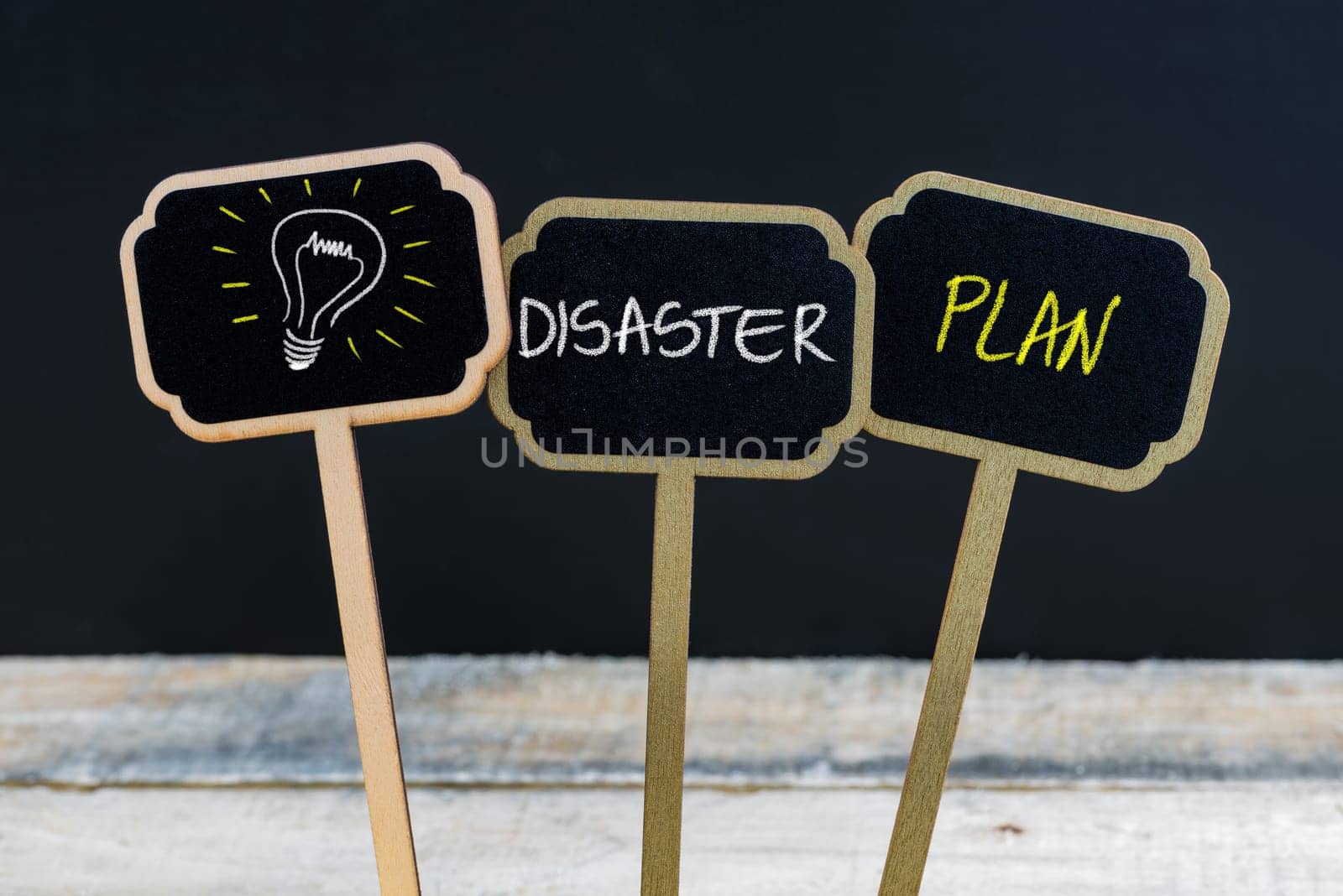 Concept message DISASTER PLAN and light bulb as symbol for idea written with chalk on wooden mini blackboard labels, defocused chalkboard and wood table in background