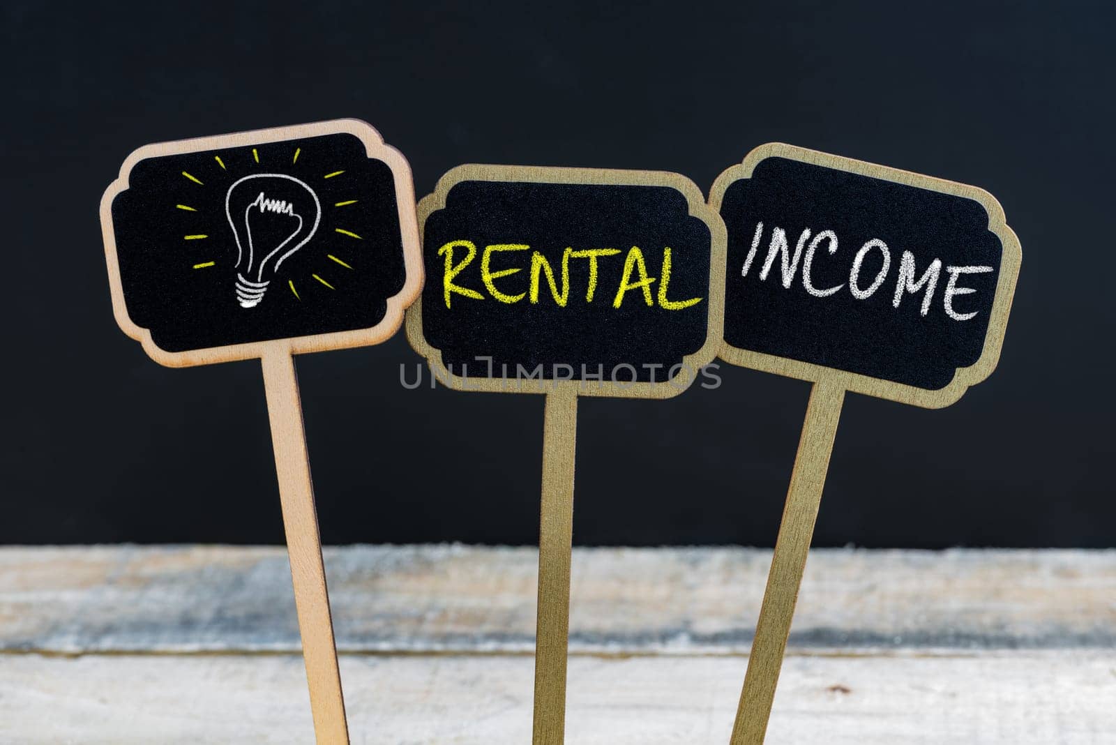 Concept message RENTAL INCOME and light bulb as symbol for idea written with chalk on wooden mini blackboard labels, defocused chalkboard and wood table in background