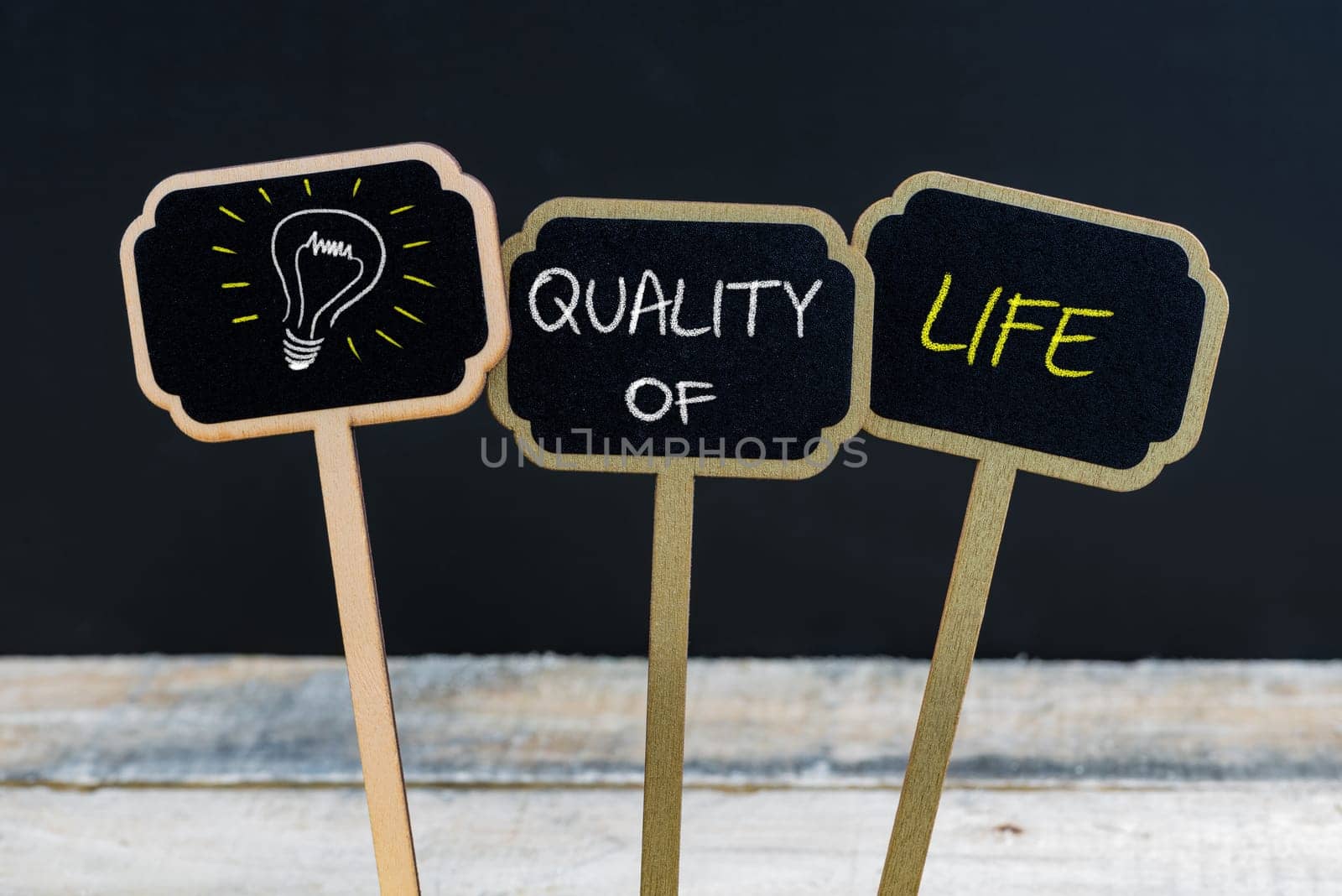 Concept message QUALITY OF LIFE and light bulb as symbol for idea written with chalk on wooden mini blackboard labels, defocused chalkboard and wood table in background