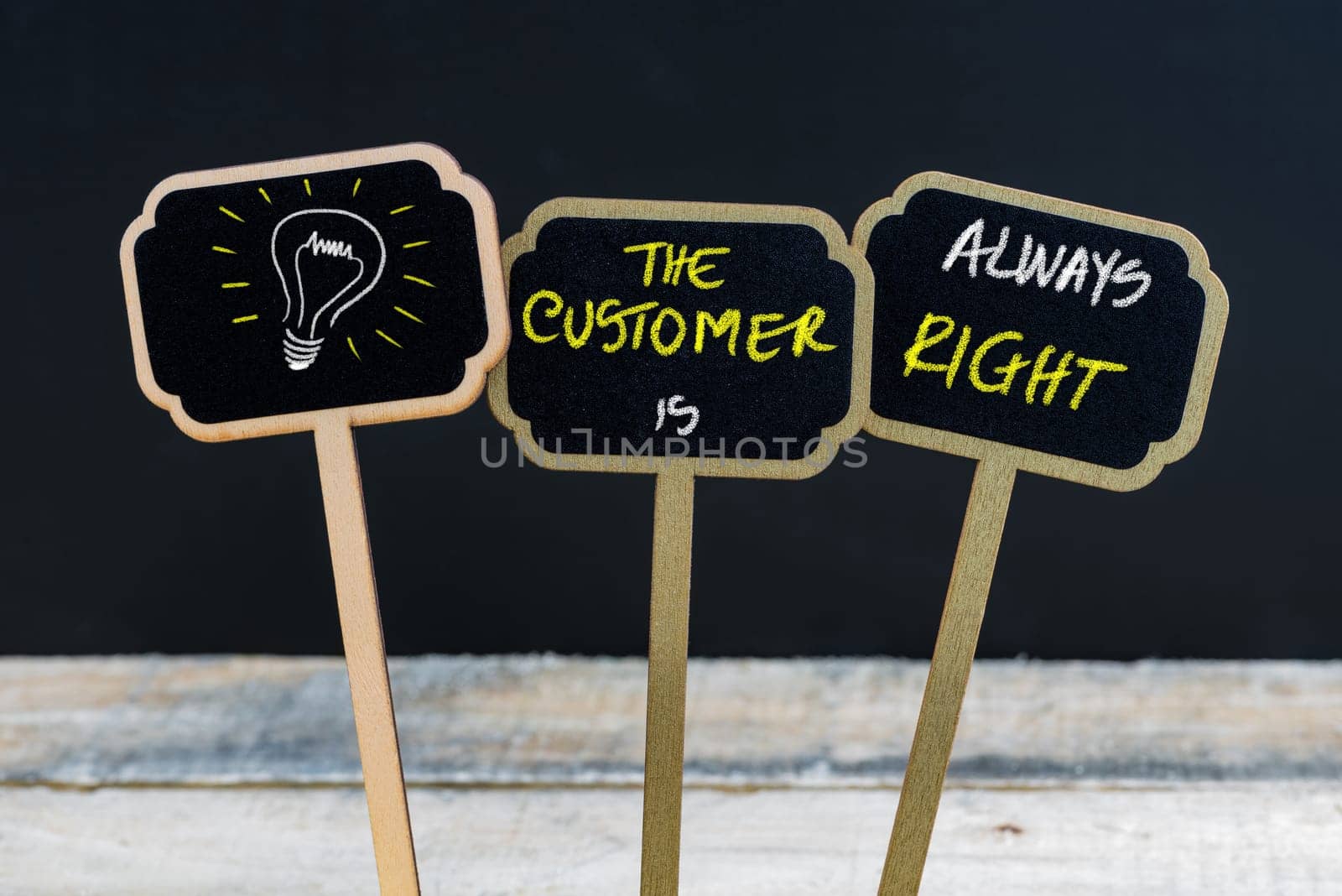 Concept message THE CUSTOMER IS ALWAYS RIGHT and light bulb as symbol for idea written with chalk on wooden mini blackboard labels, defocused chalkboard and wood table in background