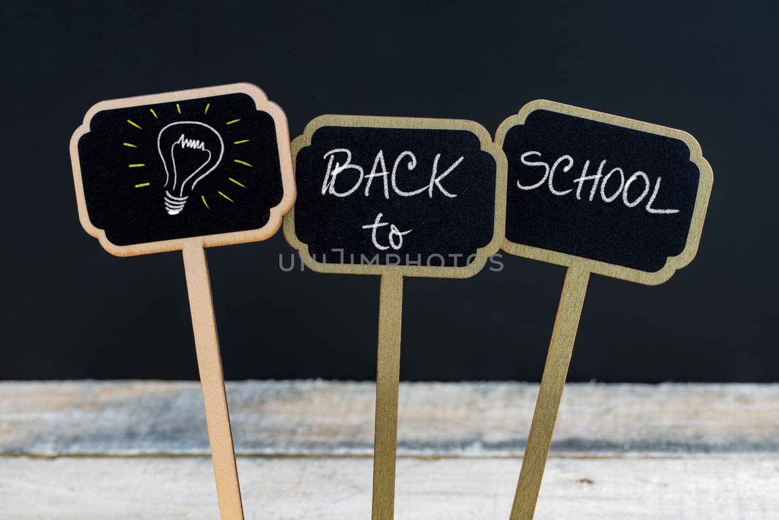 Concept message BACK TO SCHOOL and light bulb as symbol for idea written with chalk on wooden mini blackboard labels, defocused chalkboard and wood table in background