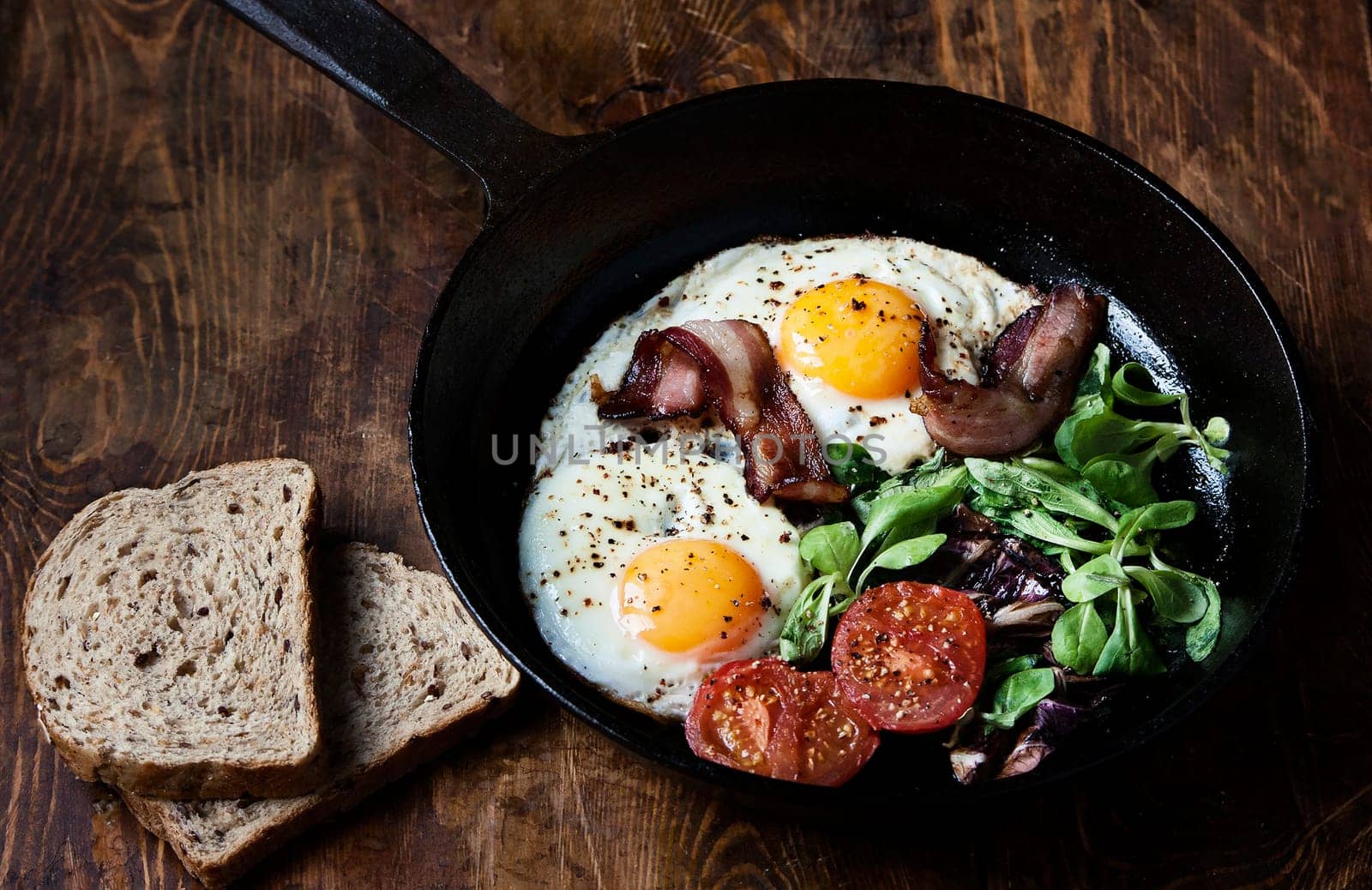 Breakfast set. Pan of fried eggs with bacon, fresh tomato, sage and bread on dark serving board over black wooden background, top view, copy space