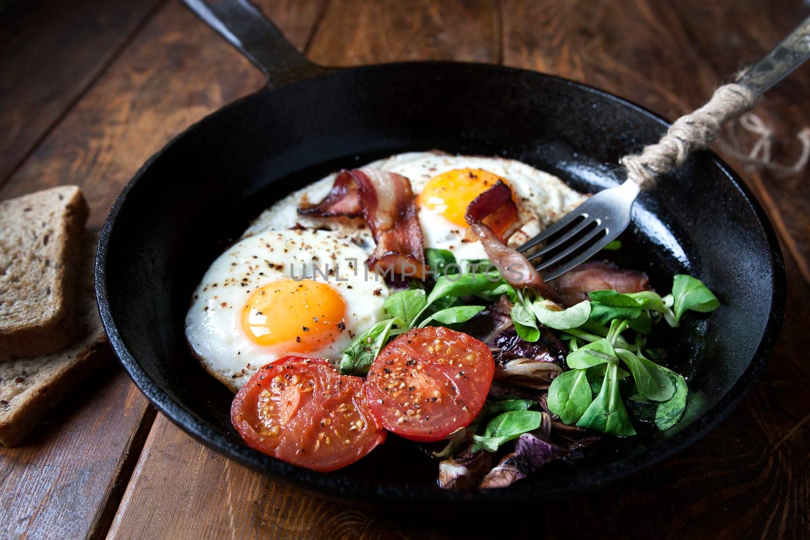 Breakfast set. Pan of fried eggs with bacon, fresh tomato, sage and bread on dark serving board over black wooden background, top view, copy space