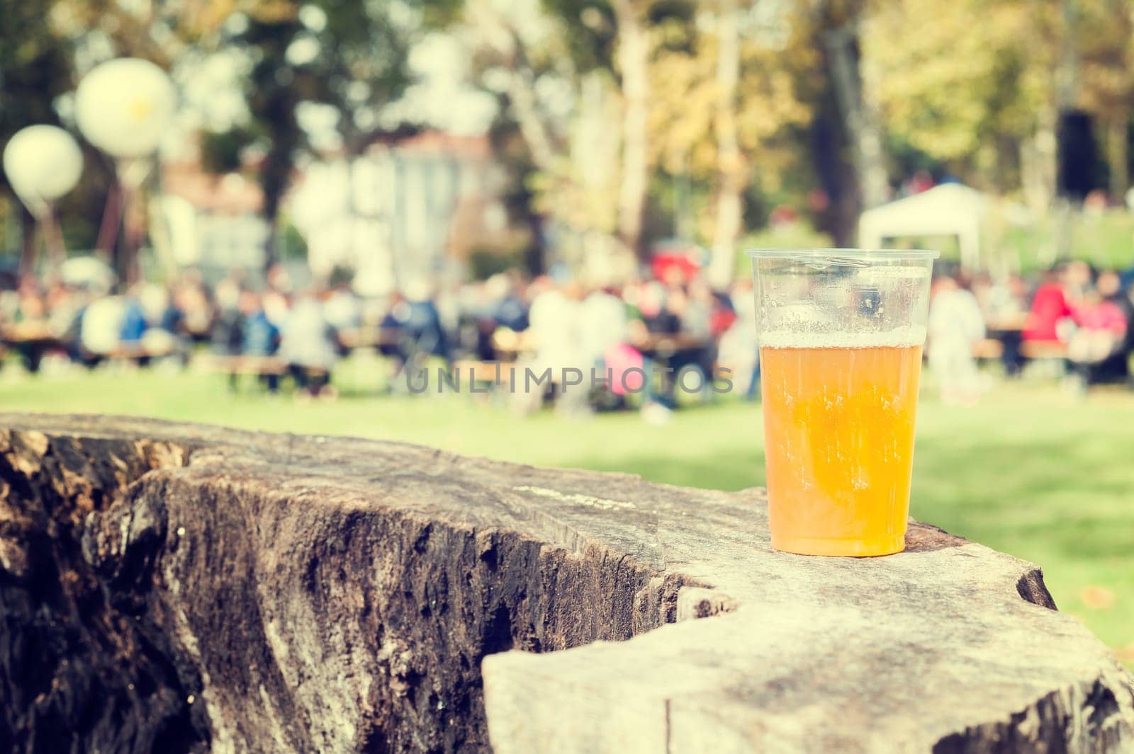Plastic cup of beer on a tree trunk. In the background, blurred people participating in a village feast. Copy space. Vintage style photo.