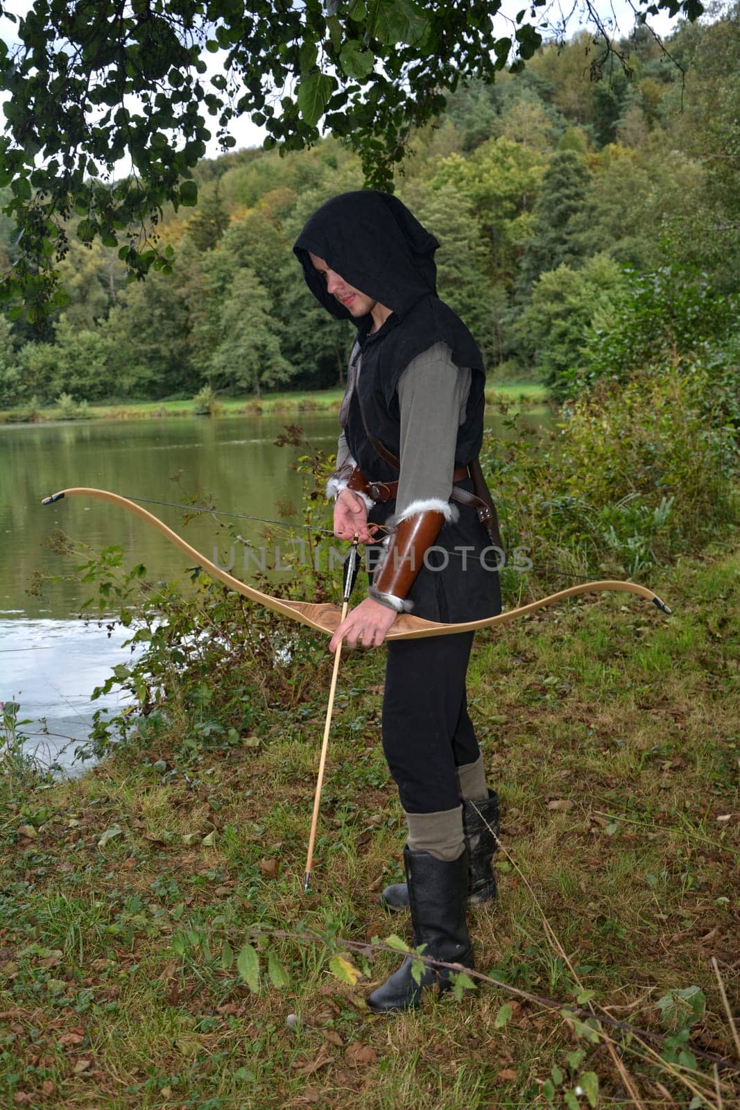 Young medieval archer with black hood stands at the lake with a tense bow and arrow