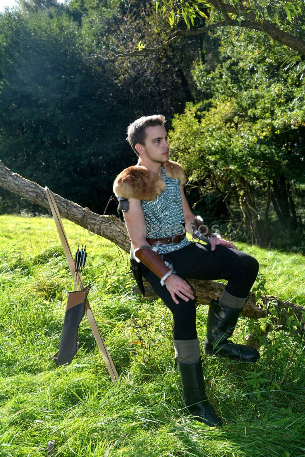 Young medieval dressed man,sitting on branch,hands on legs and looking to the side,bow and arrow stands next to it