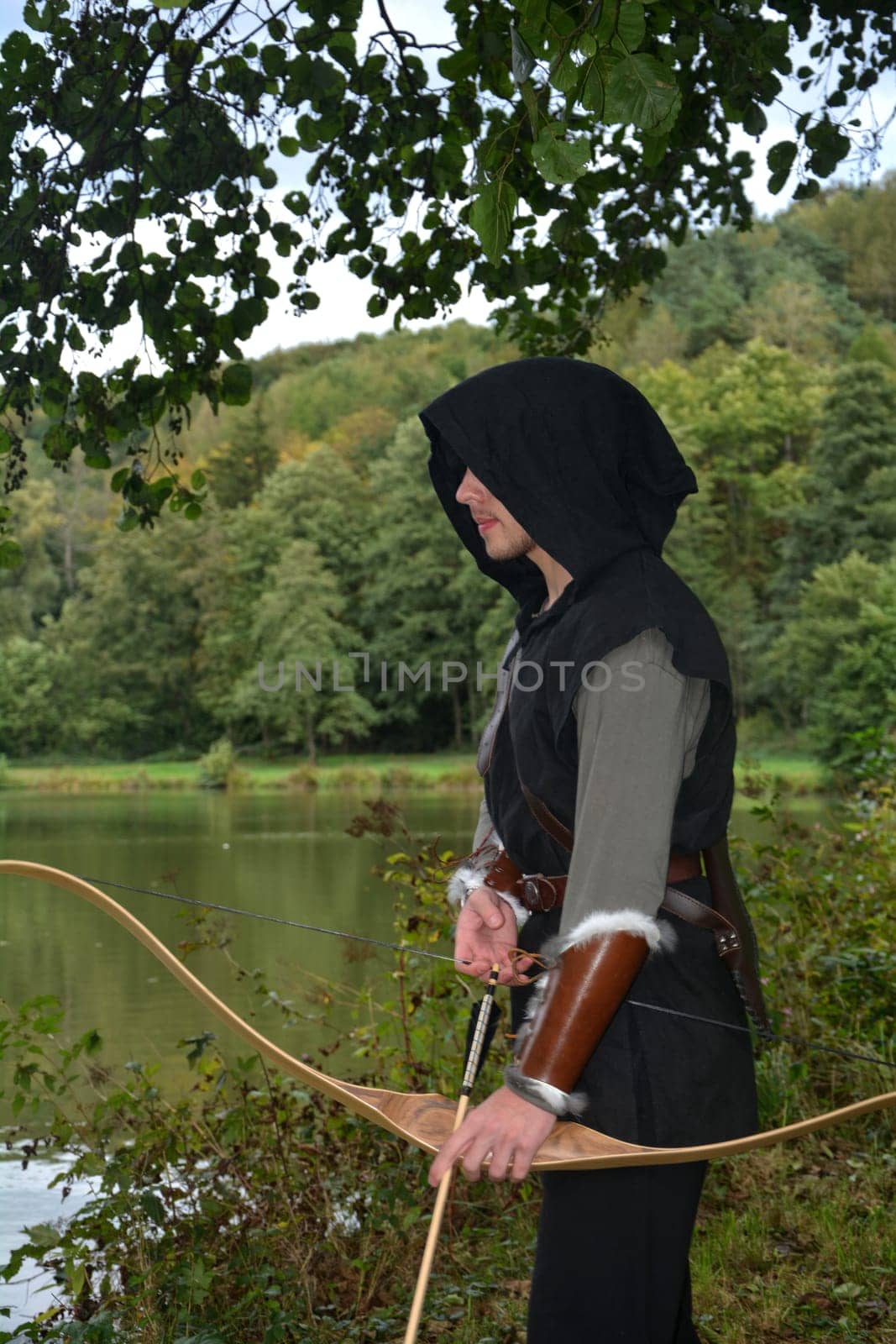Young medieval archer with black hood stands at the lake with a tense bow and arrow