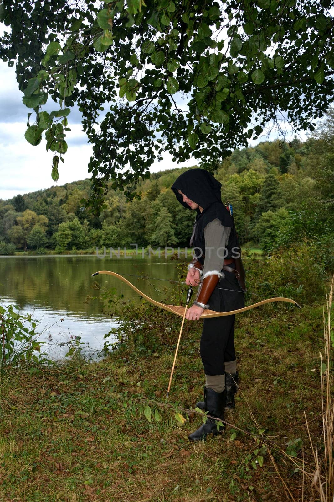 Young medieval archer with black hood stands at the lake with a tense bow and arrow