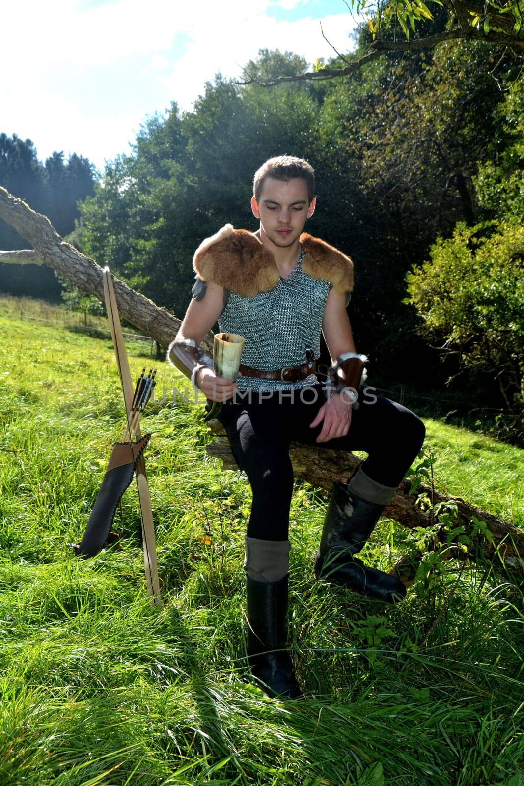 Young medieval archer with chain mail sits on branch in nature in the sunlight,drinking horn in hand,bow and arrow stands next to it