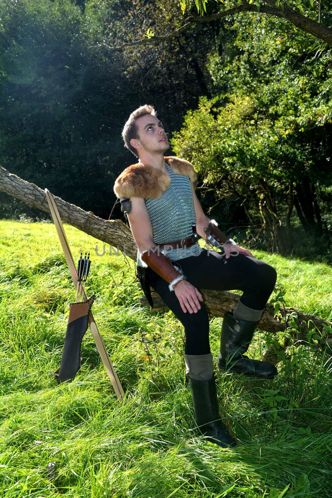 Young man dressed in medieval clothes,sits on branch,hands on legs and looks up,bow and arrow stands next to it