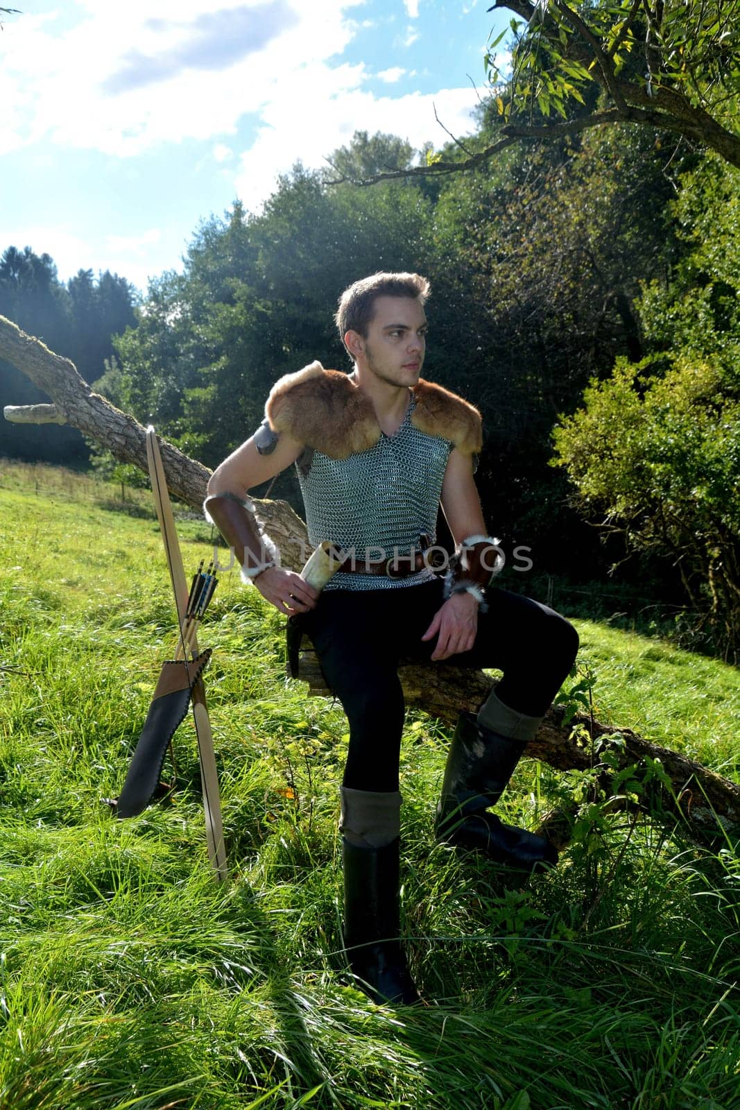 Young medieval archer with chain mail sits on branch in nature in the sunlight,bow and arrow stands next to it