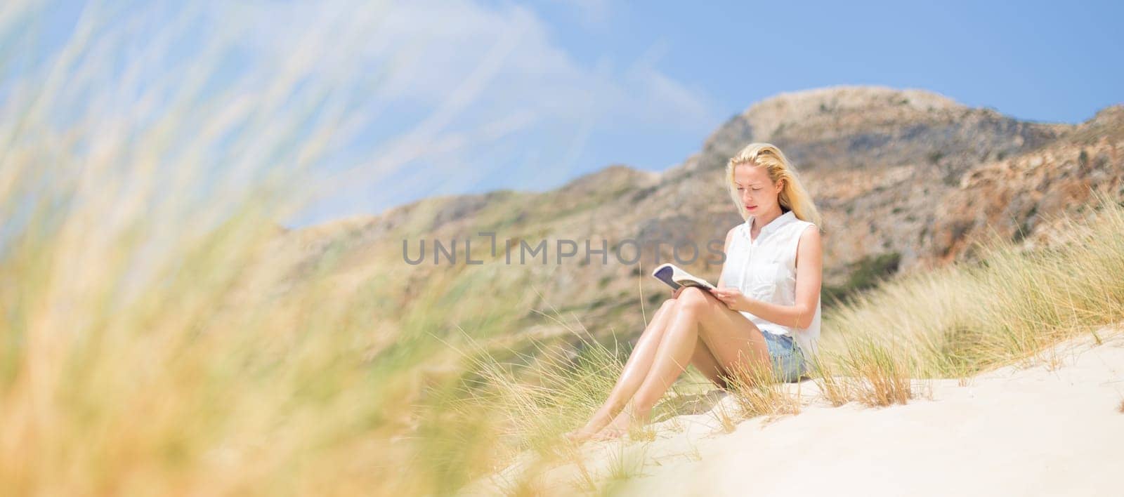 Relaxed woman enjoying sun, freedom and good book an beautiful sandy beach of Balos in Greece. Young lady reading, feeling free and relaxed. Vacations, freedom, happiness, enjoyment and well being.