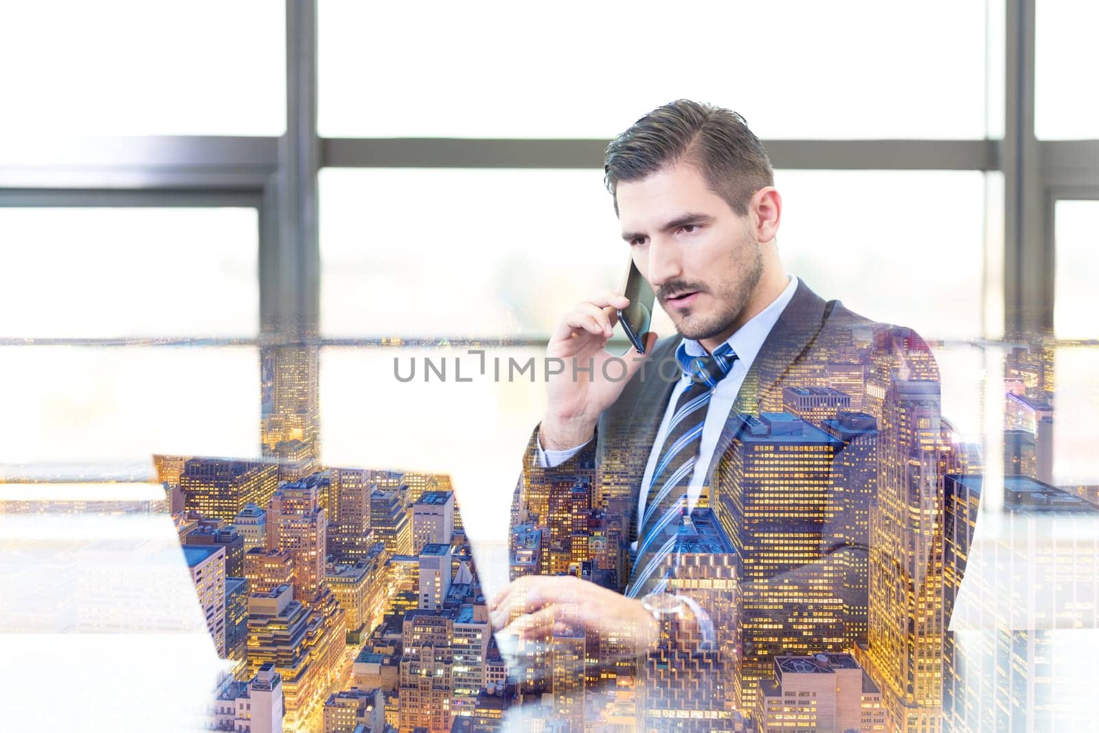 Portrait of successful corporate businessman in modern office focused on data on his laptop computer while talking on mobile phone. New Your city lights reflection in window. Business concept.