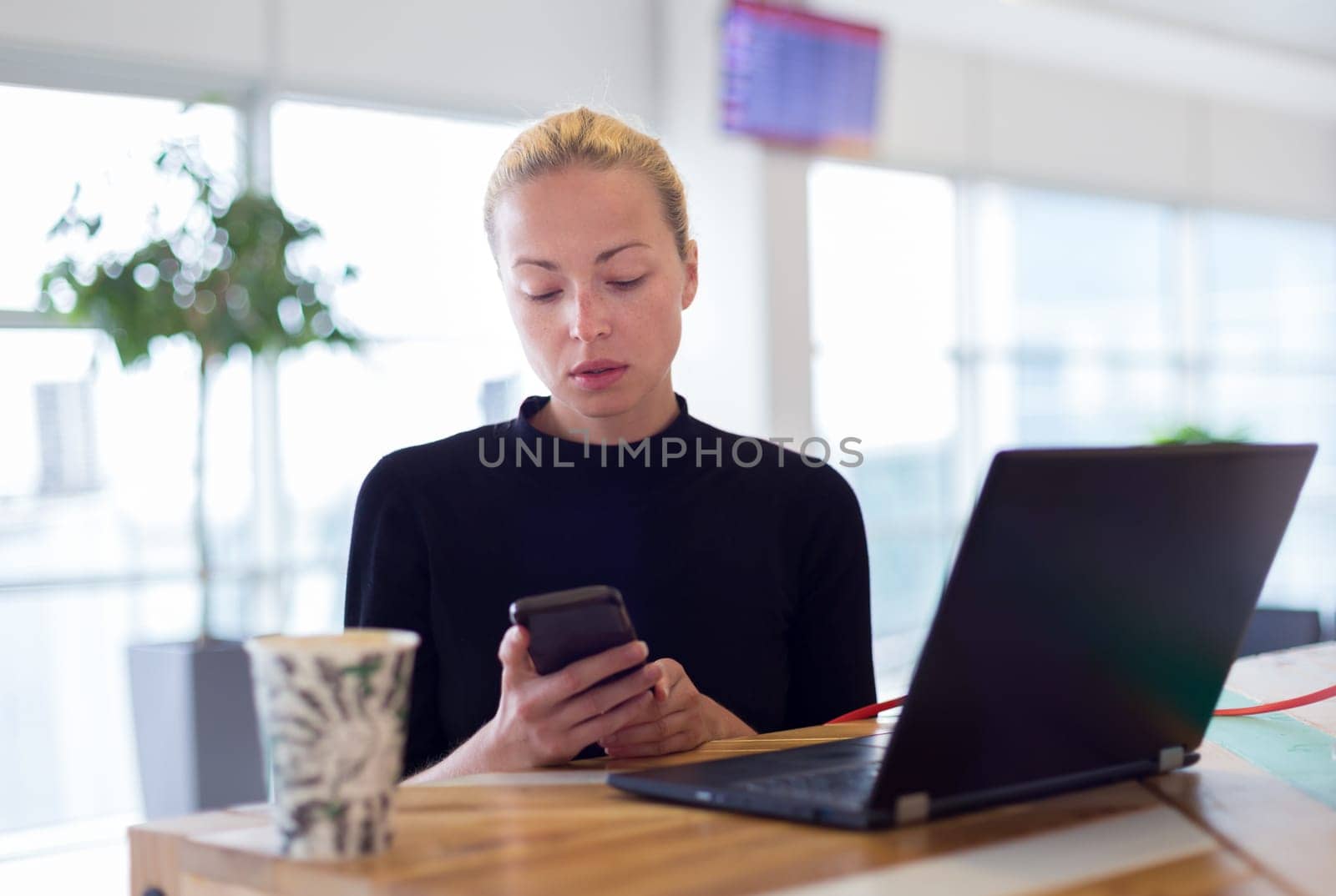 Female freelancer remotely working on the go using multiple devices on a desk at airport coffee shop while traveling. Contemporary entrepreneurial lifestyle.